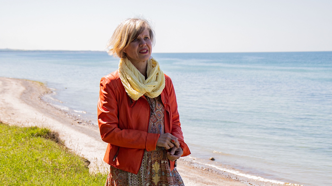 Frau in orangefarbener Lederjacke mit blonden Haaren und gelben Tuch vor der Ostsee und dem Strand im Hintergrund