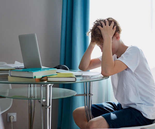 Junge sitzt verzweifelt mit beiden Händen die Haare raufend und niedergebeugten Kopf am Schreibtisch mit Computer und Schulsachen
