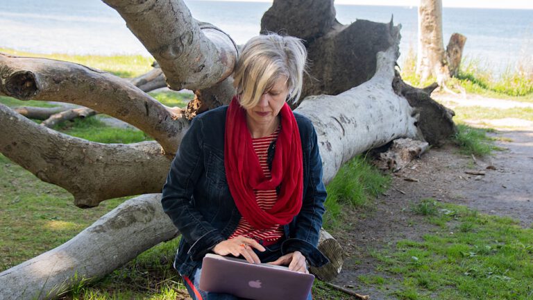 blonde Frau mit Jeansjacke und rotem Tuch sitzt auf einem Baumstamm mit geöffnetem Laptop auf dem Schoß