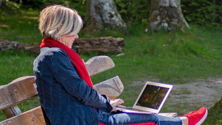 blonde Frau in Jeansjacke mit rotem Tuch sitzt mit beiden Beinen auf der Bank im Wals mit einem geöffneten Laptop auf dem Schoß