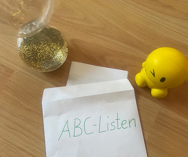 Umschlag mit der Aufschrift ABC-Listen, indem eine Liste hineingesteckt wird, Sanduhr mit goldenen Sand symbolisiert die Zeit, Smiley steht daneben und symbolisiert den Erfolg der ABC-Listen für das Lernen