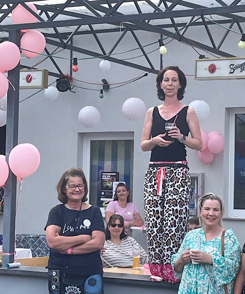 Frau steht auf dem Tisch, davor stehen rechts und links zwei blonde Frauen, rosa und weiße Luftballons sind darum herum