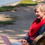 Frau in Jeansjacke und rotem Tuch sitzt an der Steilküste mit der Ostsee im Hintergrund auf der Bank und hat eine Laptop auf dem Schoß