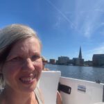 Rückblick und volle Fahrt voraus; blonde Frau im E-Boot mit der Silhouette der Hansestadt Rostock im Hintergrund