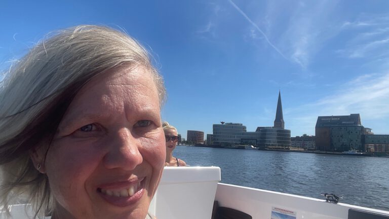 Rückblick und volle Fahrt voraus; blonde Frau im E-Boot mit der Silhouette der Hansestadt Rostock im Hintergrund