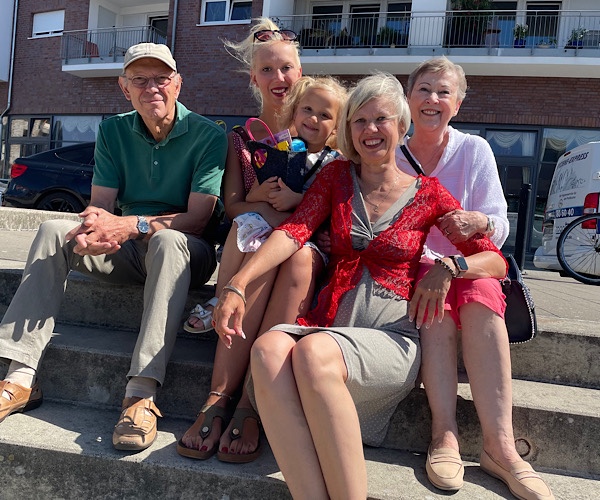 Familie in vier Generationen mit blonder Frau im Sommerkleid, erwachsener Tochter, Enkelin und Eltern sitzen auf den Hafenterassen im Rostocker Stadthafen mit einem Gebäude auf der Holzhalbinsel im Hintergrund