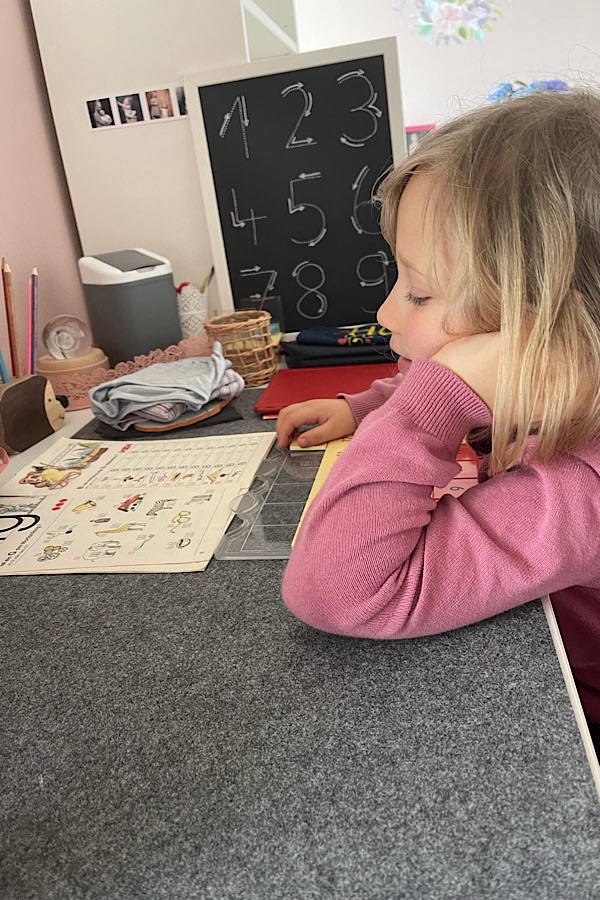 blondes Mädchen sitzt am Tisch, stützt den Kopf auf die Hand, der Oberarm liegt auf der Tischplatte, das Kind bearbeitet Aufgaben mit dem LÜK-Kasten