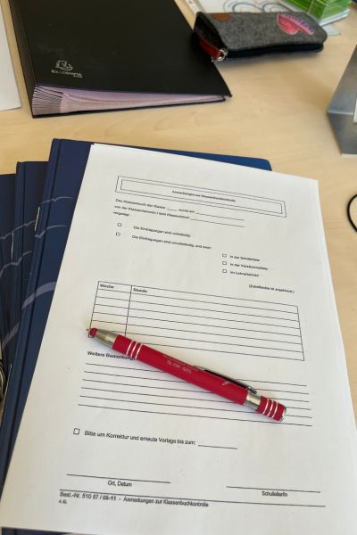 12 von 12 im Mai - Stapel Klassenbücher mit einem Formblatt für Rückmeldungen der Klassenbuchkontrolle, roter Kugelschreiber liegen auf dem Schreibtisch