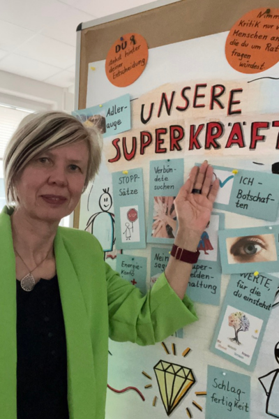 Monatsrückblick April 2025: blonde Frau im grünen Blazer zeigt auf das Plakat "Unsere Superkräfte"