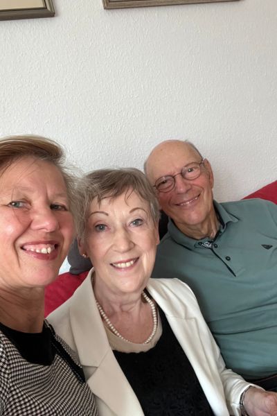 Monatsrückblick 2025: Frau Ende 50 im schwarz weiß karierten Kleid, Frau Ende 70 im weißen Blaser und Mann ohne Haare mit Brille im grünen Shirt sitzen dicht zusammen auf dem Sofa