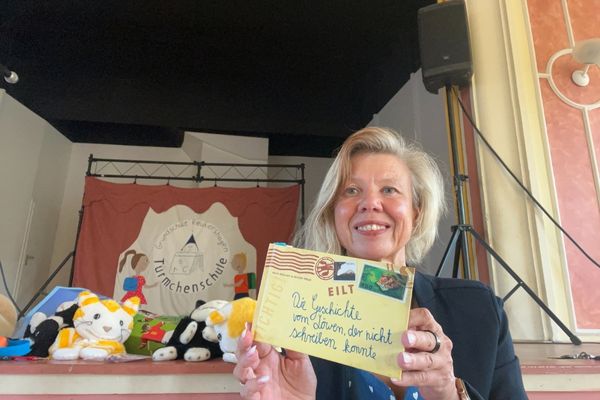 Monatsrückblick 2025: blonde Frau in dunkelblauer Jacke und blauem Kleid mit weißen Punkten hält ein Buch in der Hand; im Hintergrund ist eine Bühne mit den Puppen Mimi und Mo und dem Logo der Türmchenschule