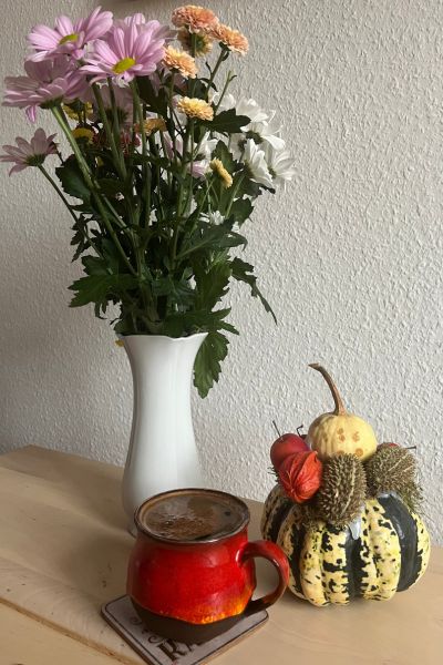 12 von 12 im Oktober 2025; bunte Herbstblumen in einer weißen Vase, ein dekorierter Kürbis und eine Tasse Kaffee auf einem holzfarbenen Tisch vor weißer Wand
