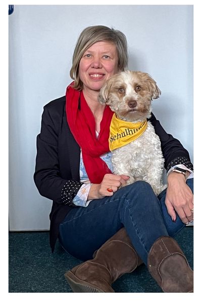 Frau mit blonden Haaren in Jeanshosen und blauer Jacke sitzt vor einer weißen Tür mit einem Bolonka Zwetna auf dem Schoß, der Hund hat ein gelbes Tuch mit der Aufschrift Schulhund umgebunden