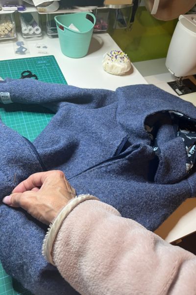 blauer Walkoverall liegt auf dem Nähtisch, eine Hand hält eine Nähnadel