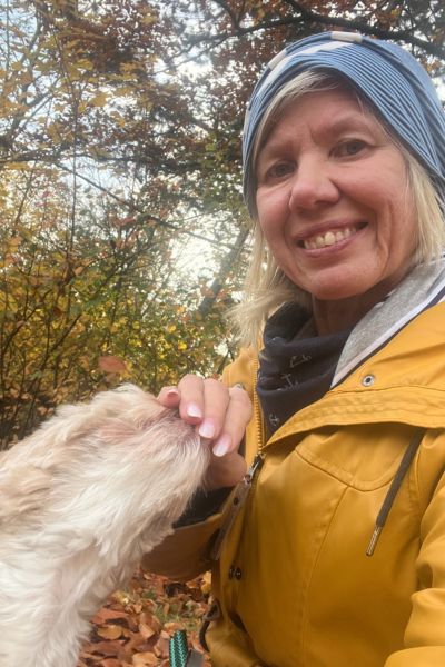 Frau im senffarbenen Regenmantel und blauer Mütze freut sich zum Bolonka Zwetna, der ihre Hand küsst