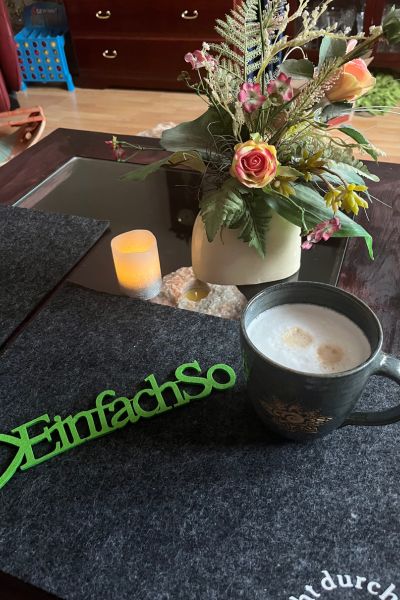 Tisch mit Blumen und einer Tasse Milchkaffee, auf dem Tisch liegt der Schriftzug "Einfach so"