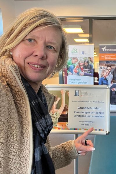 blonde Frau in hellbrauner Jacke zeigt auf ein Schild mit der Auffschrift: Grundschulklar - Erwartungen der Schule an die Schulanfänger