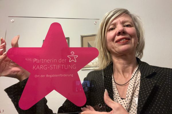 Monatsrückblick Dezember 2025 Lernen Familie Glaube; Frau hält ein Schild mit einem pinken Stern und der Aufschrift: Ort der Begabtenförderung und Partner der Karg Stiftung