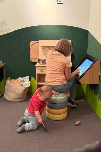 Kind lernt trotz Üben nicht: blondes achtjähriges Kind sitzt auf bunten Stapelsteinen in der Spielecke und tippt  am Monitor. Davor hockt ein zweijähriger Junge im roten Pulli.