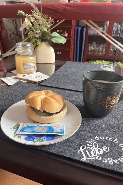12 von 12 im Mai 2026: Wohnzimmertisch mit einer Tasche Kaffee und einem Teller mit einem Brötchen