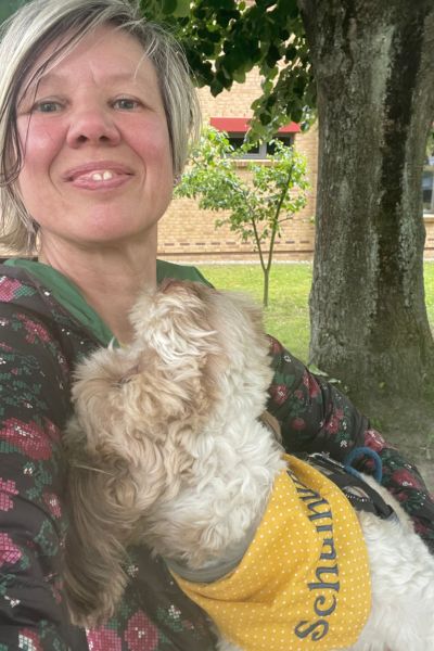 Schulhund Addi: Bolonka Zwetna mit einem gelben Tuch und der Aufschrift Schulhund schaut eine blonde Frau an