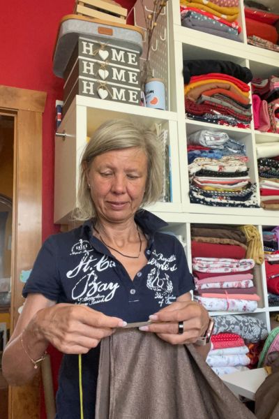 12 von 12 im August 2026: Frau Mitte fünfzig steht vor einem Regal voller Stoff und kürzt eine Hose