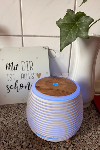 Anwendungsarten ätherischer Öle: ein diffuser blau leuchtend steht auf einer Fläche, im Hintergrund steht ein Schild mit dem Spruch "Mit dir ist alles schön"