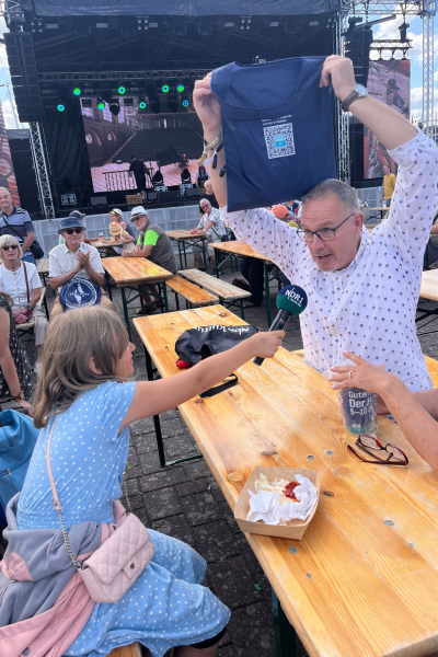 Monatsrückblick August 2026: Kind und Reporter vor der Bühne hält dem Moderator das Mikrofon