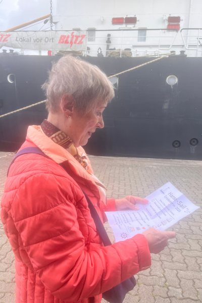 Monatsrückblick August 2026: ältere Frau vor einem Schiff stehend mit einem Blatt in der Hand auf der eine Hanse Sail Rallye steht