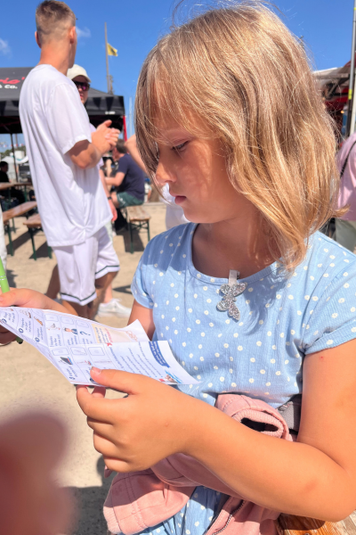 Monatsrückblick August 2026: Kind im blauen Kleid auf der Hanse Sail in Rostock hält ein Blatt mit einer Hanse Sail Rallye für Kinder in der Hand