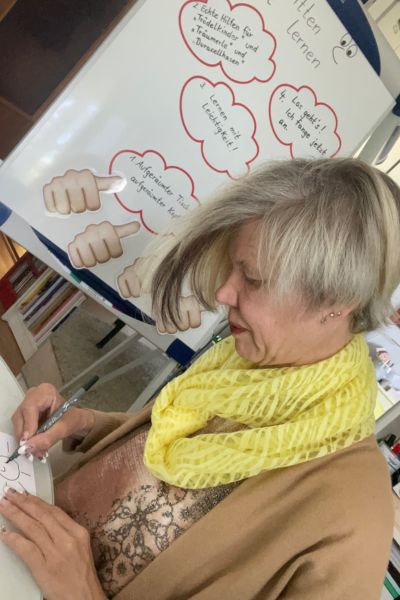 Ein Lerncoach kennt nicht immer die Lösung: Frau sitzt am Tisch und schreibt, im Hintergrund steht ein Flipchart mit Lernhinweisen